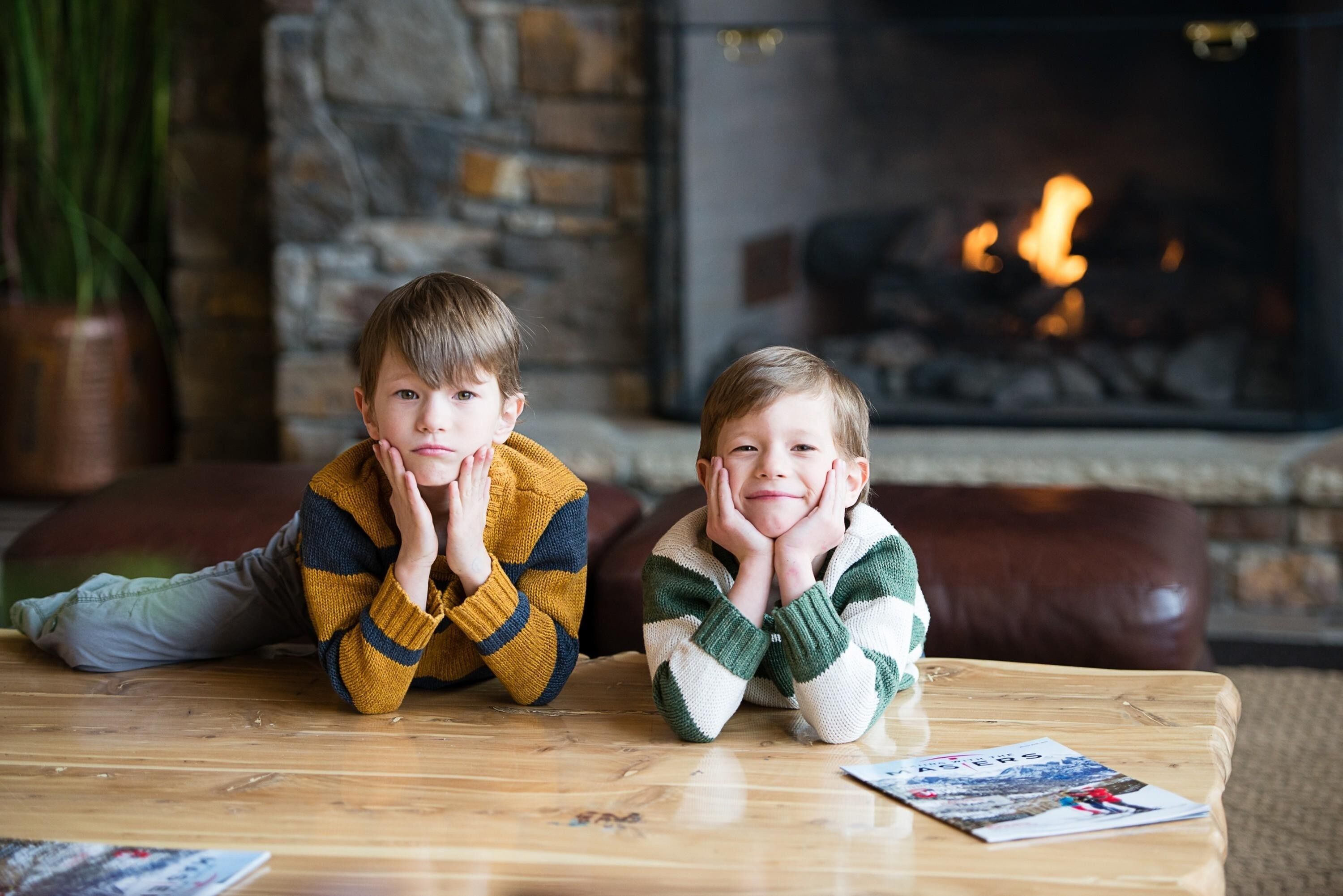 Two children sitting on a wooden coffee table in a living room with a fireplace in the background.