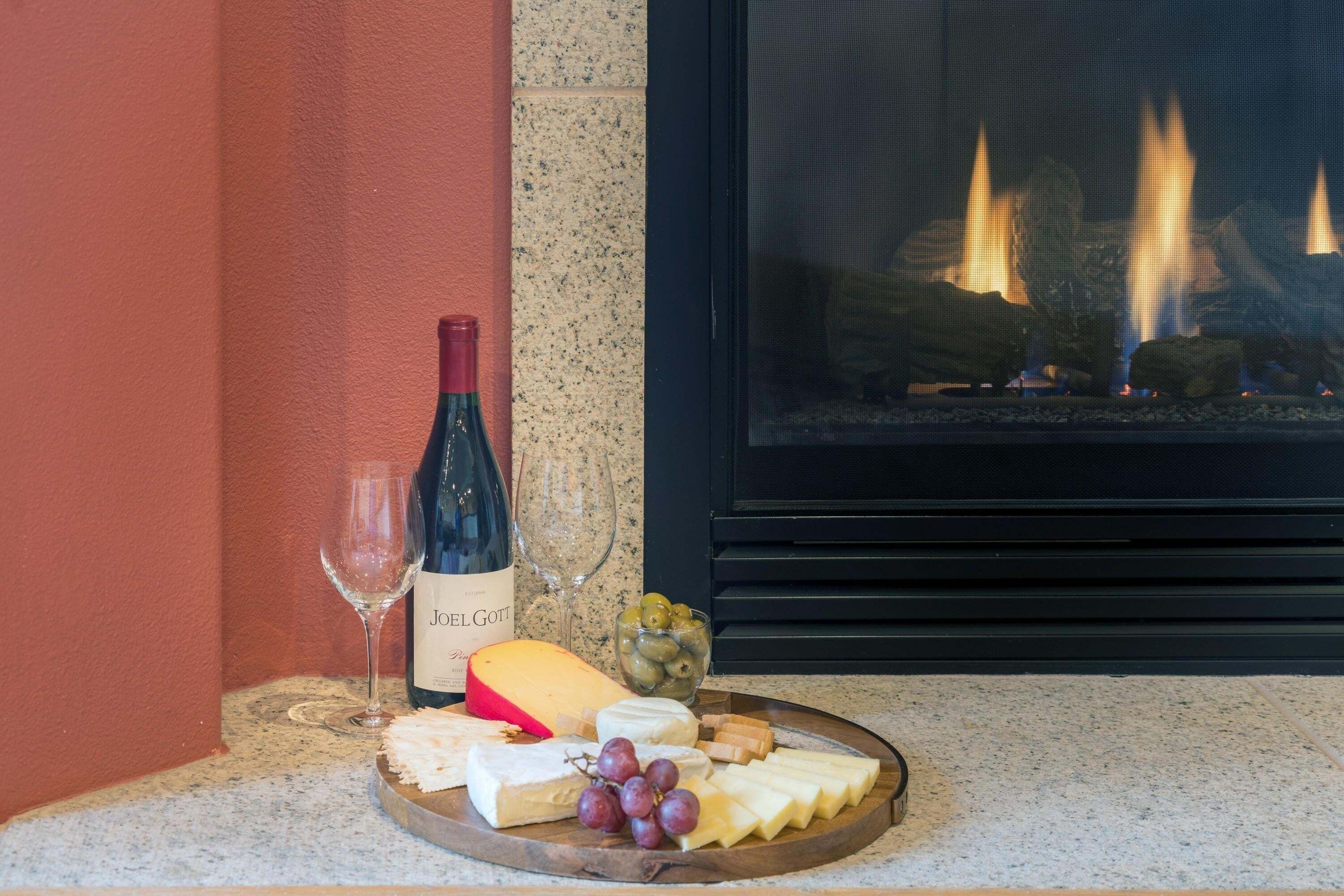 A cheese board with various cheeses, grapes, olives, and crackers, accompanied by a bottle of wine and a glass, set against a backdrop of a fireplace.
