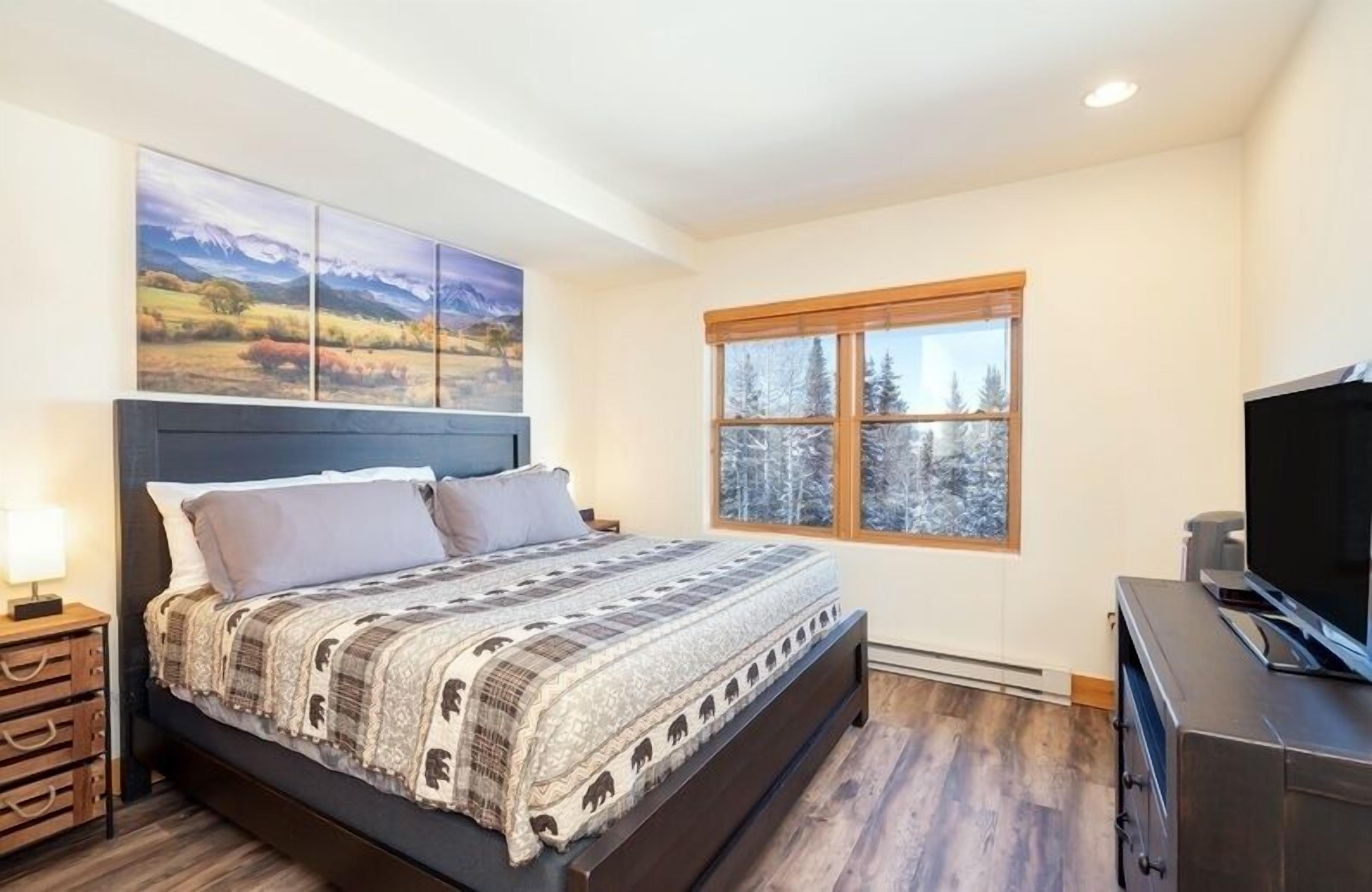 A bedroom with a large bed, a TV, a window with a view of trees, and a painting above the bed.