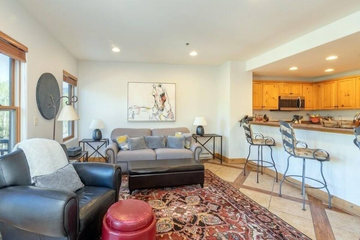 A modern living room with a sofa, ottoman, and a red stool. There is a kitchen area with wooden cabinets and a microwave.
