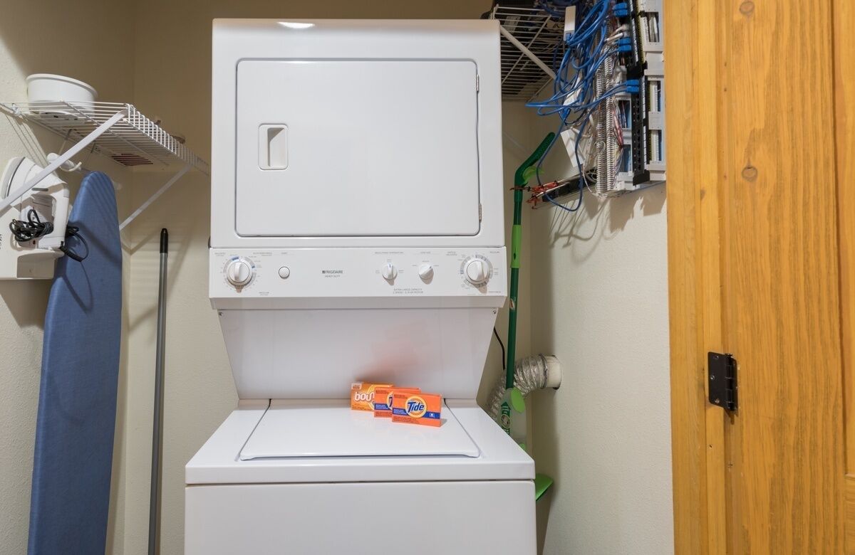 A laundry room with a stacked washer and dryer, a shelf with laundry supplies, and a wooden door.