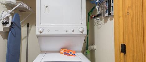 A laundry room with a stacked washer and dryer, a shelf with laundry supplies, and a wooden door.