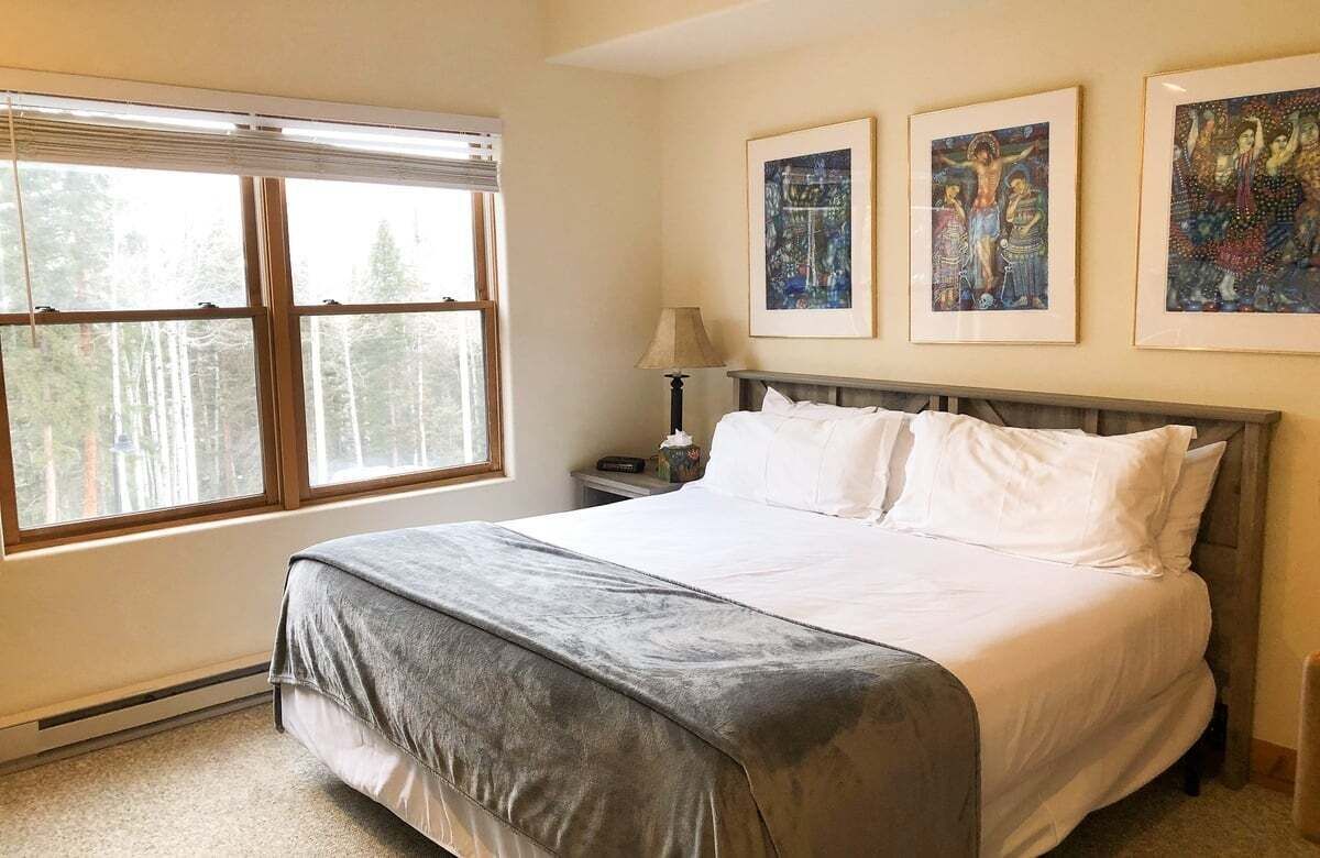 A bedroom with a large bed, a window with blinds, and three framed artworks on the wall.