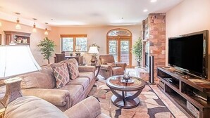 Un salon spacieux avec un grand canapé, une table basse et un téléviseur à écran plat. De nombreuses fenêtres laissent entrer la lumière naturelle et il y a une cheminée.