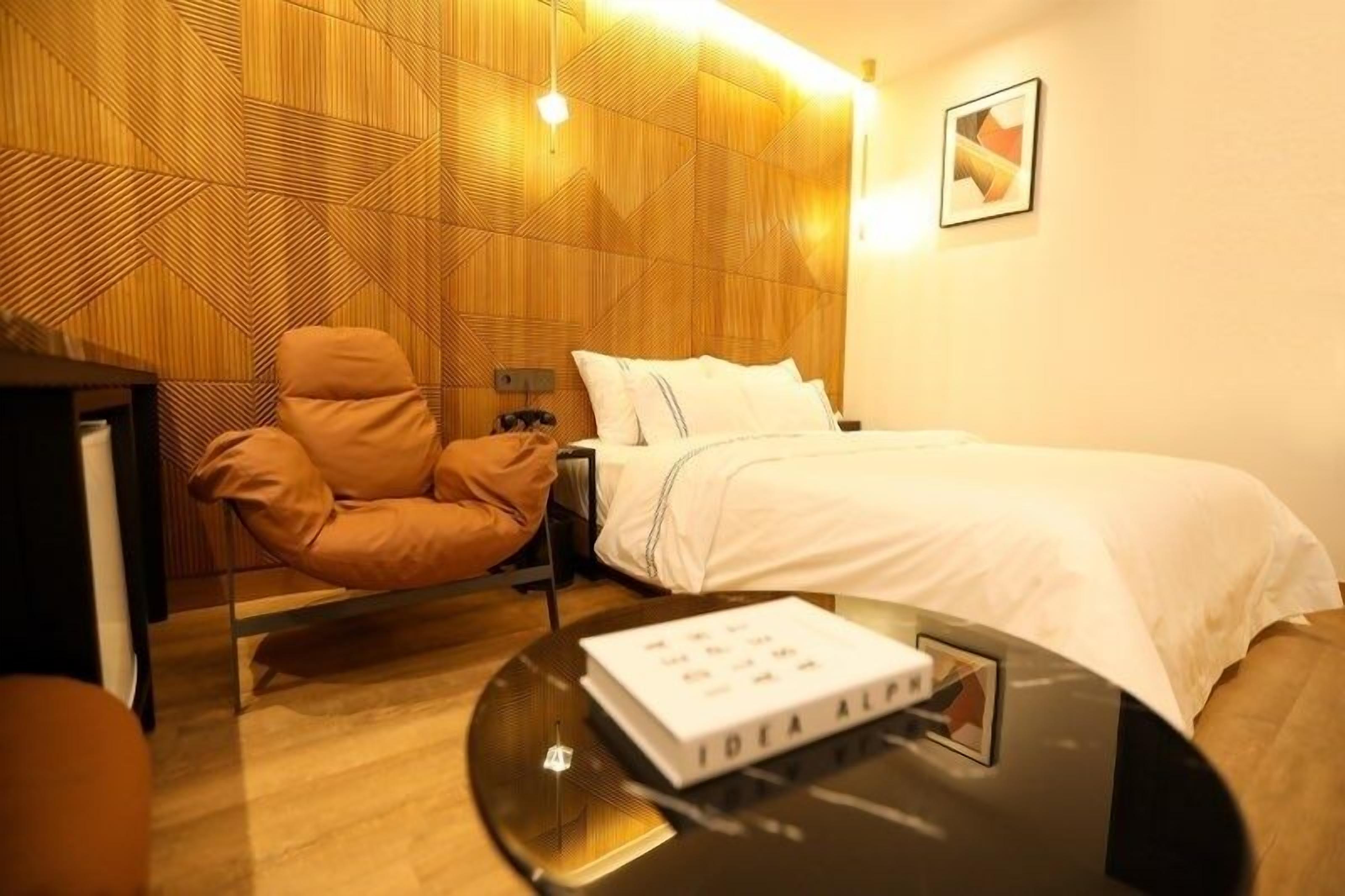 A modern hotel room with a wooden headboard, a single bed, a massage chair, and a small table with a book.