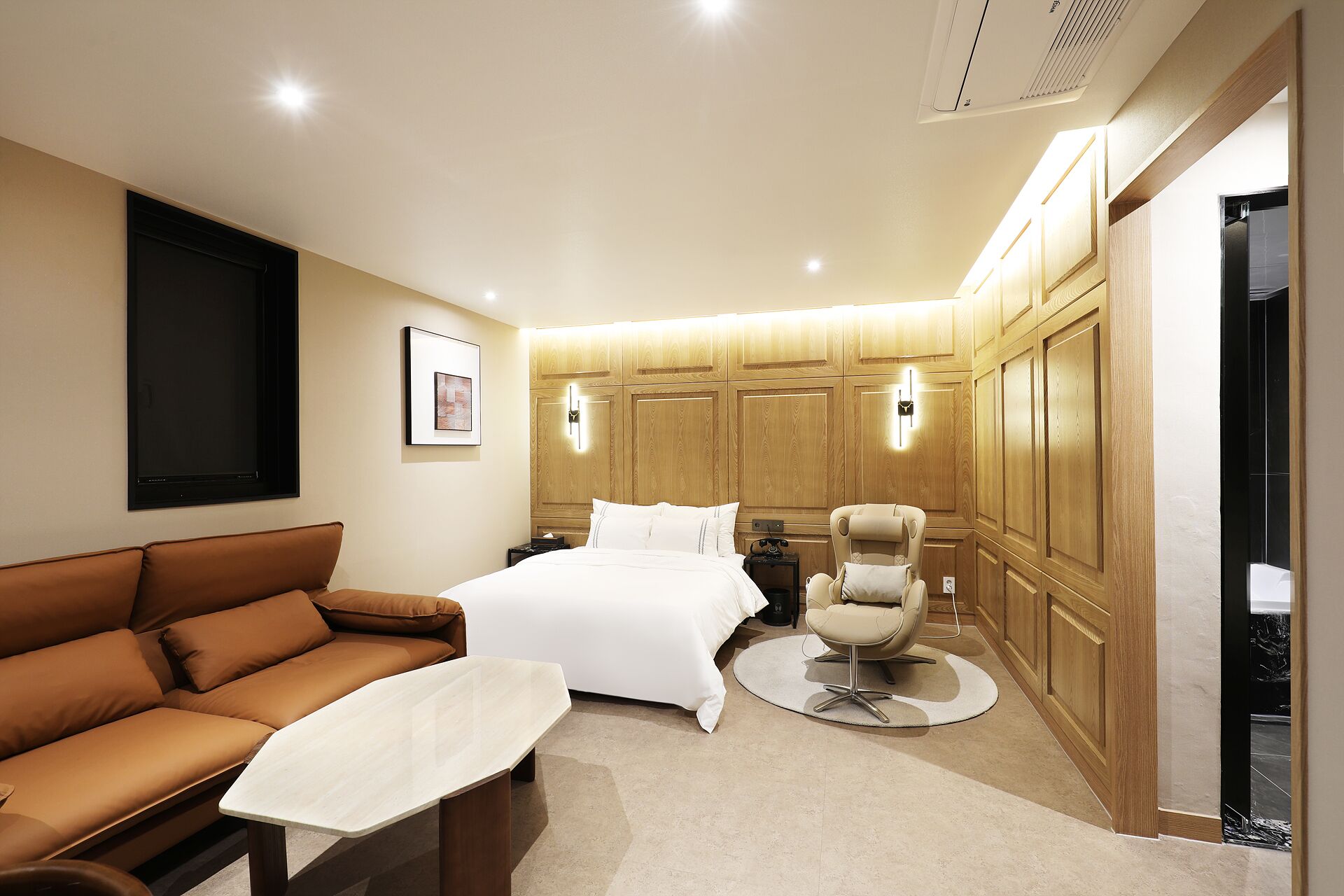 Una habitación de hotel moderna con una cama grande, un sofá de cuero marrón, una mesa de centro redonda y un armario de madera.