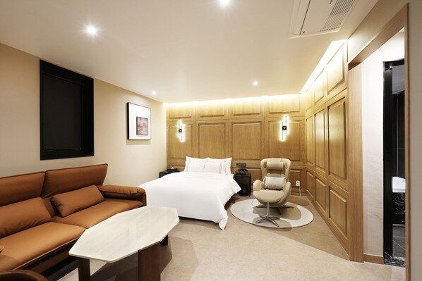 A modern hotel room with a large bed, a brown leather sofa, a round coffee table, and a wooden wardrobe.