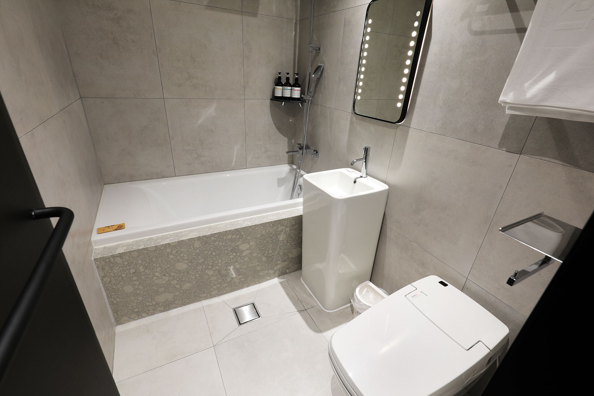 A modern bathroom with a bathtub, a sink, and a toilet. There is a shelf with toiletries and a mirror with lights.