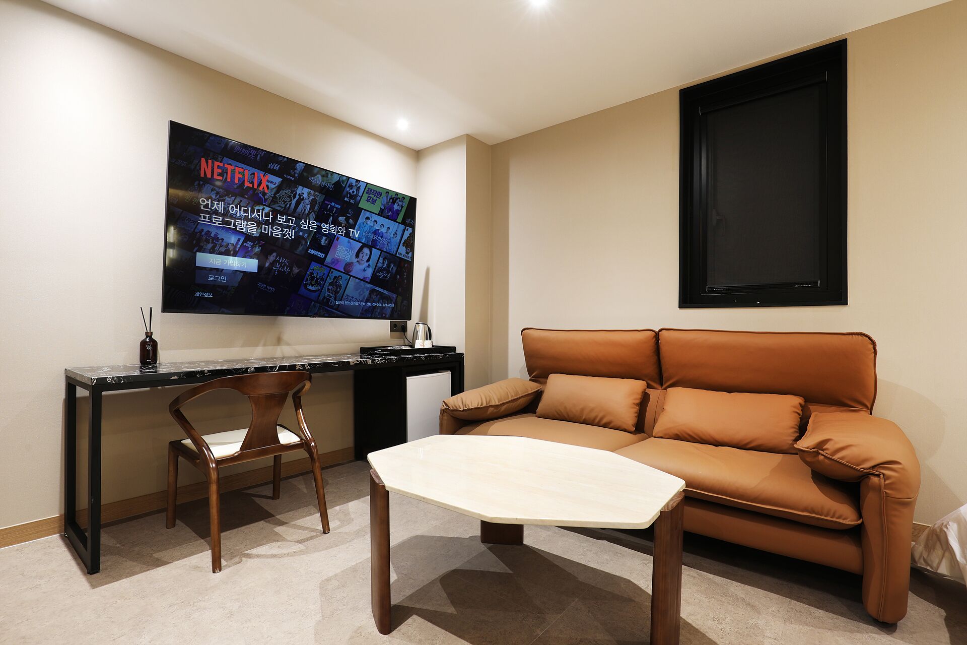 A modern living room with a large flat-screen TV mounted on the wall, a brown leather sofa, a wooden coffee table, and a wooden chair.