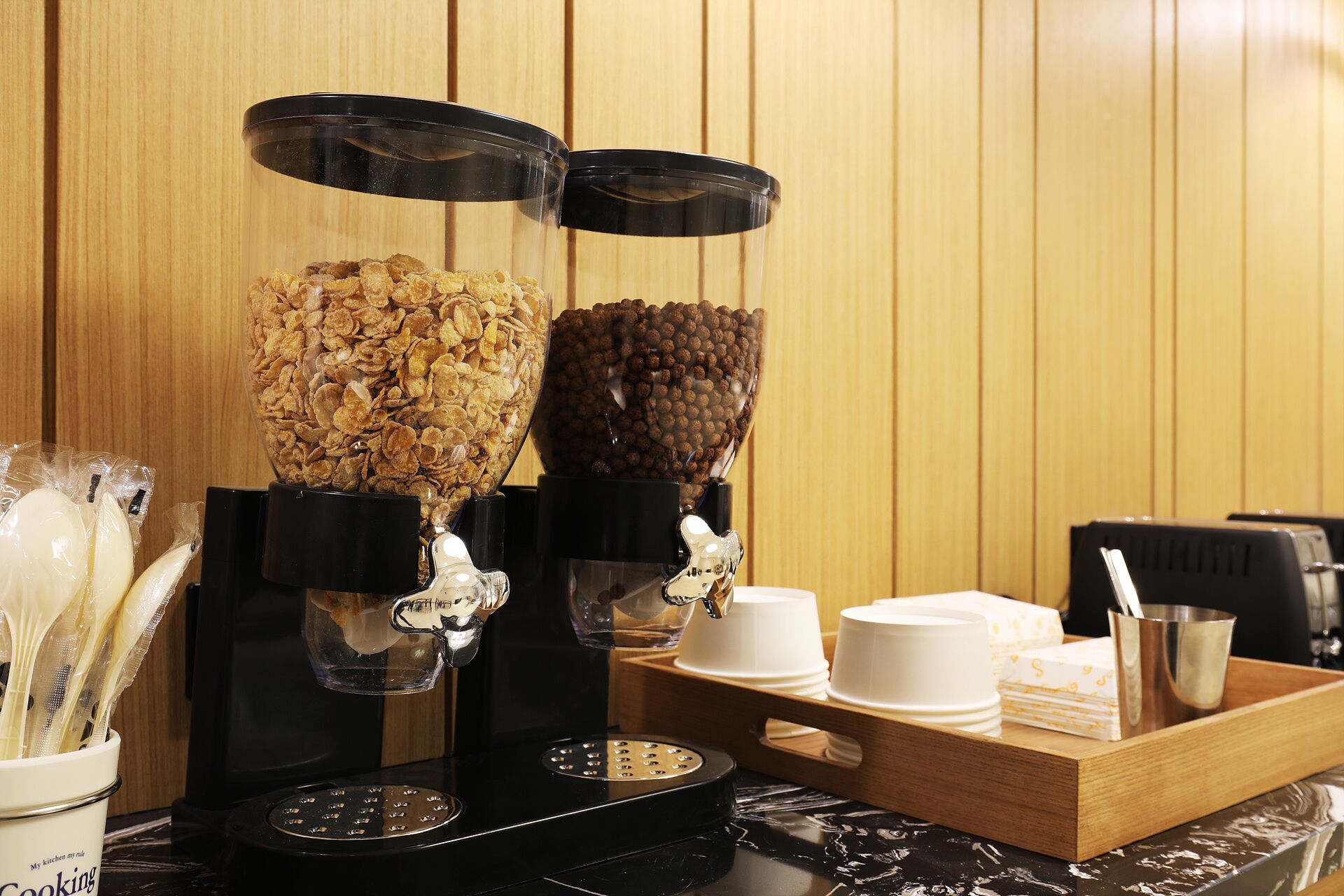 A kitchen area with two coffee dispensers, a tray with white cups and a metal pitcher, and a stove.