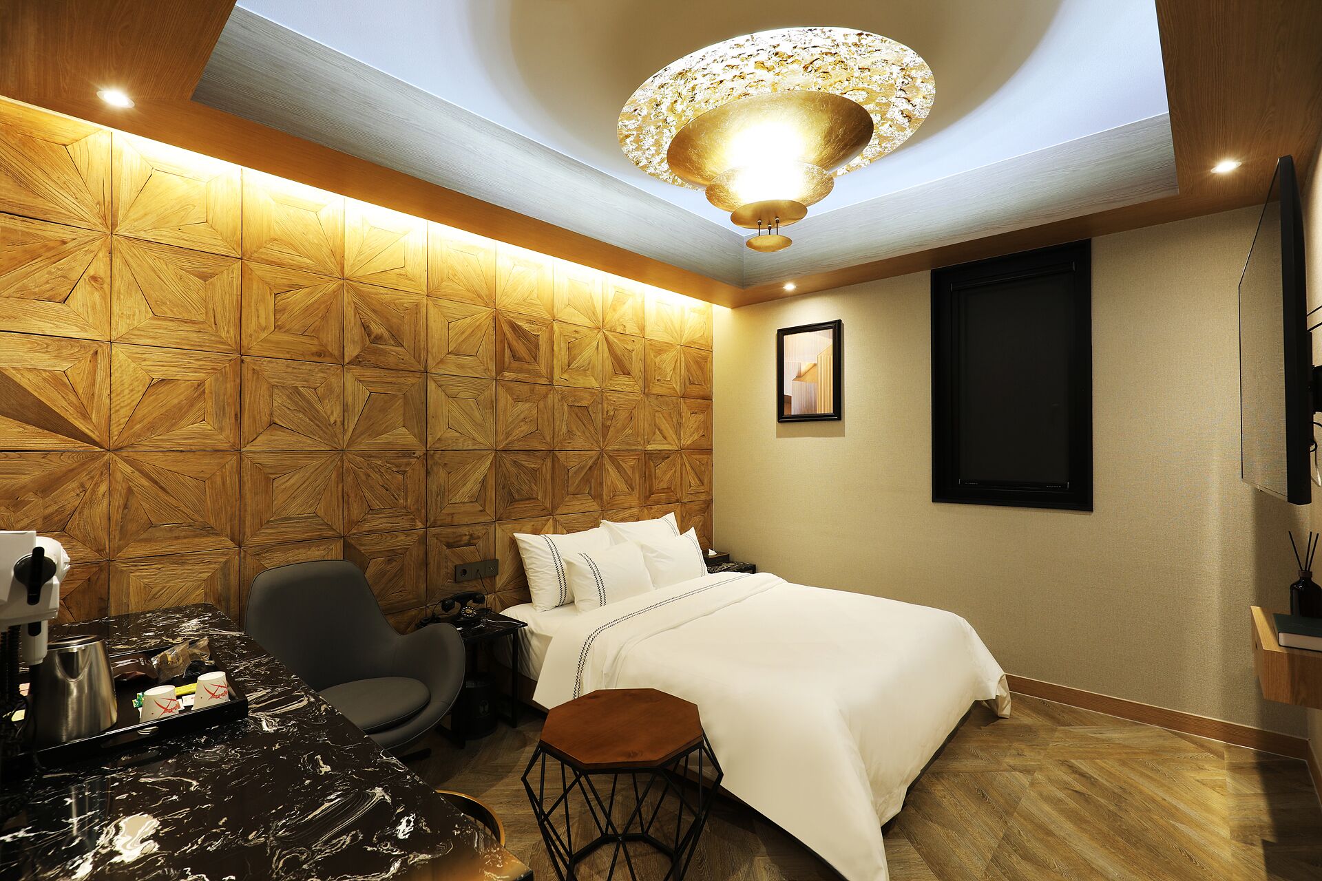 A hotel room with a geometric wooden wall, a bed with white linens, a modern chair, a small wooden side table, and a ceiling light fixture.