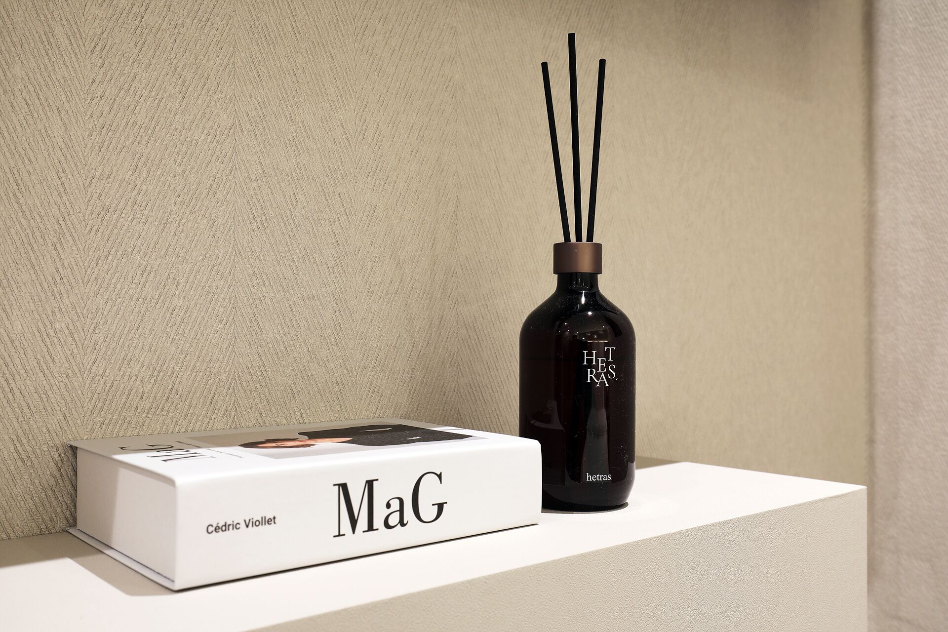 Una caja blanca etiquetada 'MaG' y una botella negra con palitos negros sobre una superficie blanca, frente a una pared con textura.