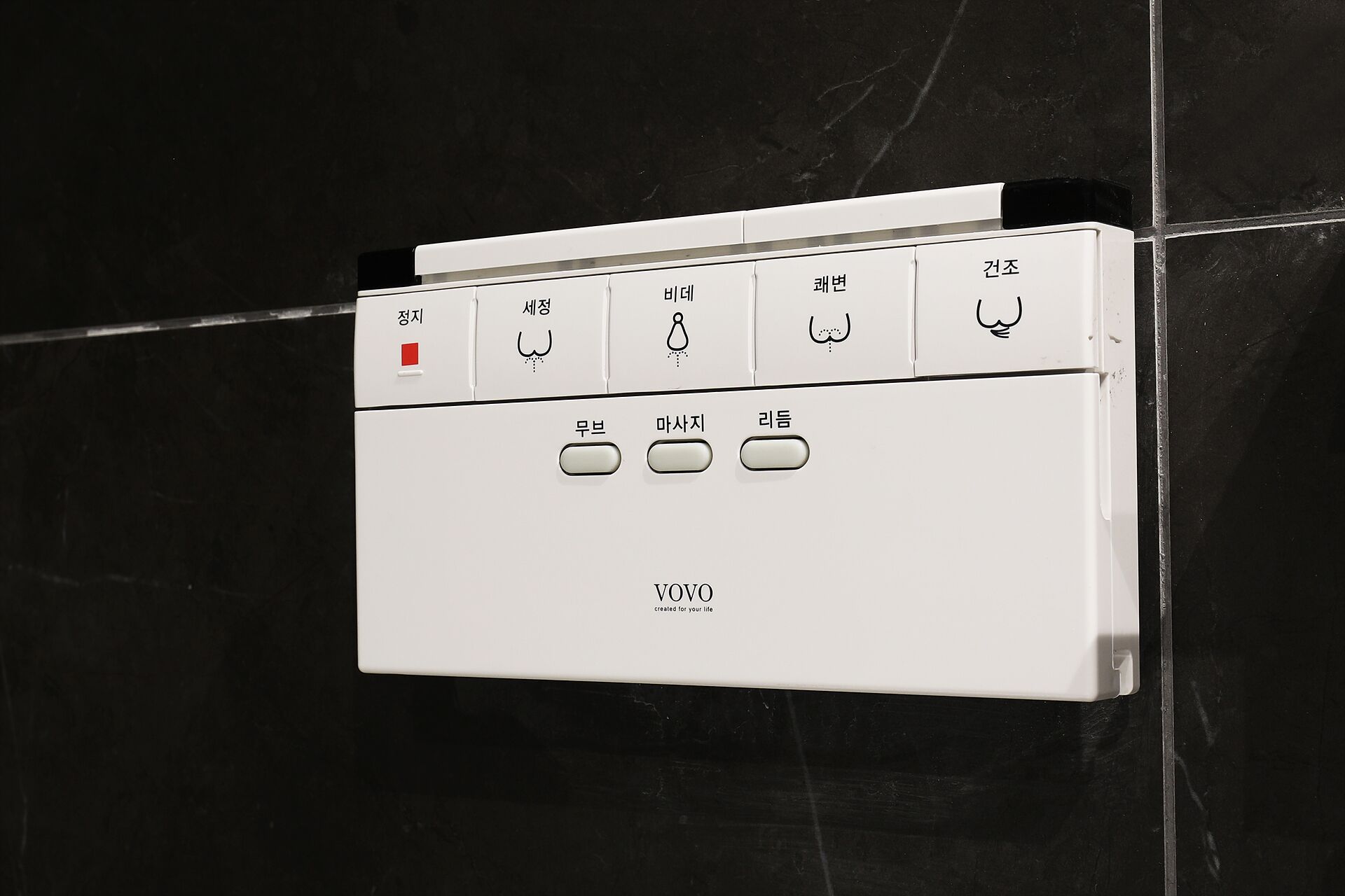 A white control panel with various buttons and labels, likely for a bathroom appliance, mounted on a black tiled wall.