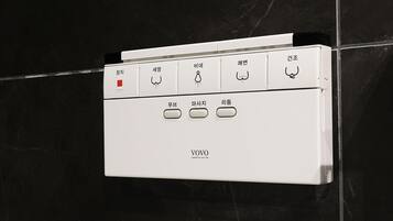 A white control panel with various buttons and labels, likely for a bathroom appliance, mounted on a black tiled wall.