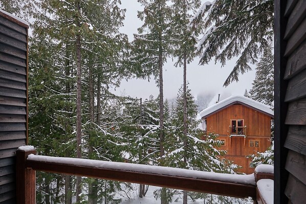 A wooden cabin with a balcony, surrounded by tall evergreen trees covered in snow.