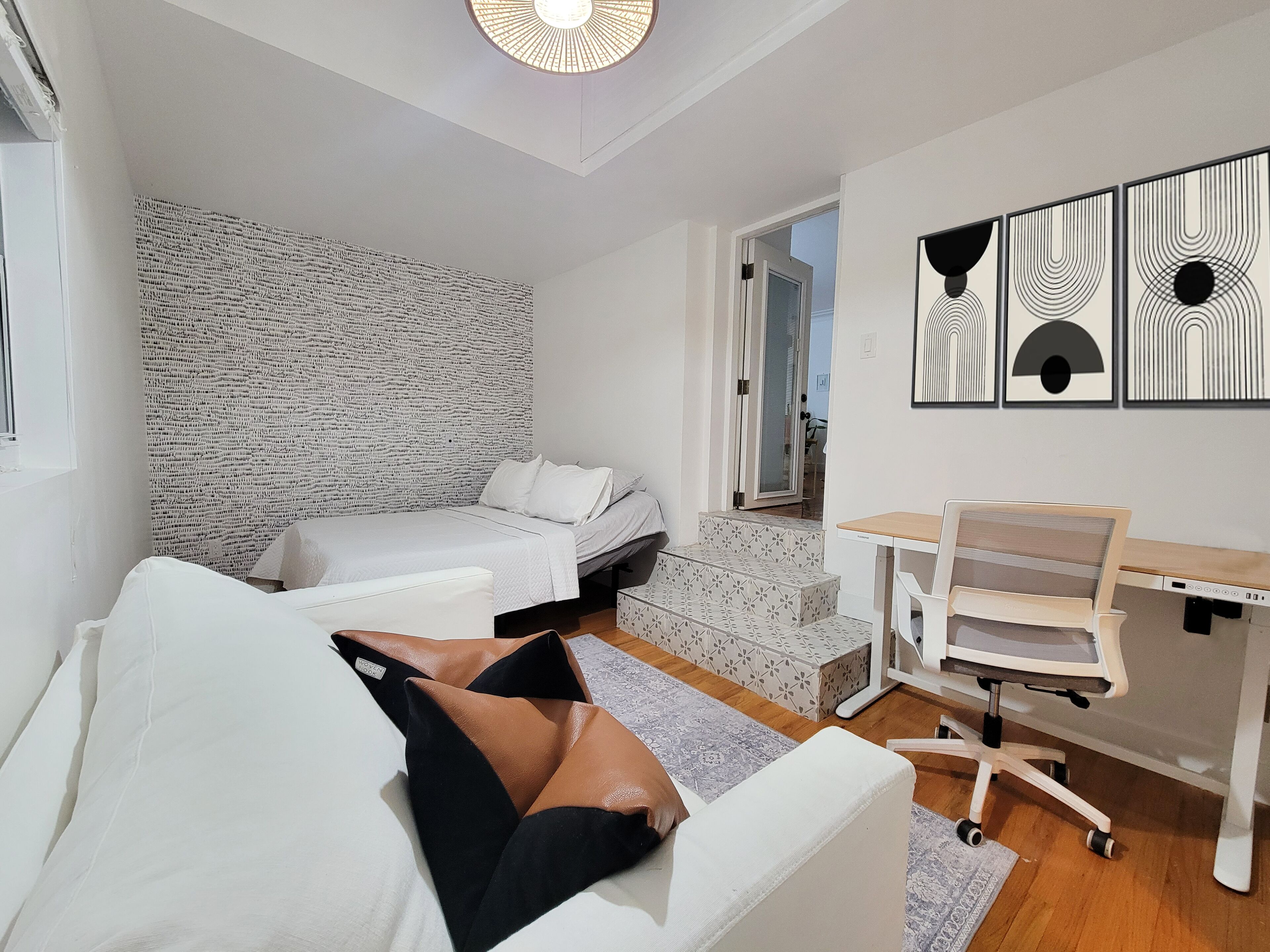 Habitación pequeña con cama, escritorio y silla. En la pared detrás de la cama se aprecia un diseño y una puerta que da a otra habitación.