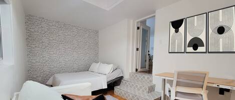 Habitación pequeña con cama, escritorio y silla. En la pared detrás de la cama se aprecia un diseño y una puerta que da a otra habitación.