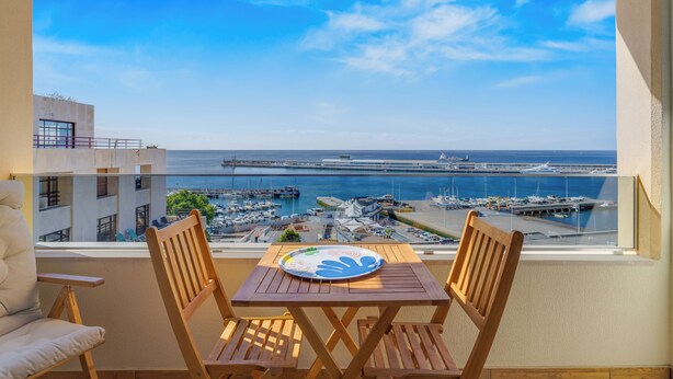 A balcony with a wooden table and two chairs, overlooking a marina with boats and a clear blue sky. - Apartamento Vista Pontinha (Funchal)