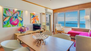 A modern hotel room with a large window offering a sea view, a dining area with a wooden table, and two vibrant paintings on the wall. - Apartamento Vista Pontinha (Funchal)