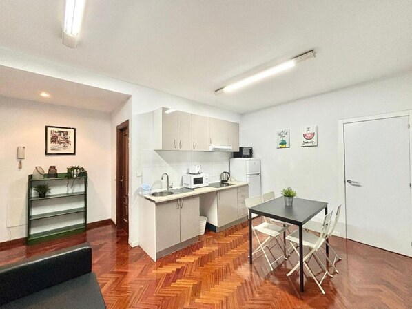 A compact kitchen with white cabinets, a microwave, a kettle, and a sink. There's a dining table with chairs and a small potted plant on the table.