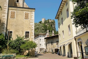 A narrow street with buildings on either side, a castle on a hill in the background, and outdoor seating areas in front of the buildings. - Muri Di Arco (Arco)