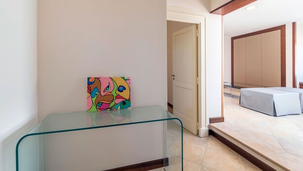 Una habitación de hotel con una mesa de vidrio, un cuadro colorido, una cama con colcha gris y suelo de madera.
