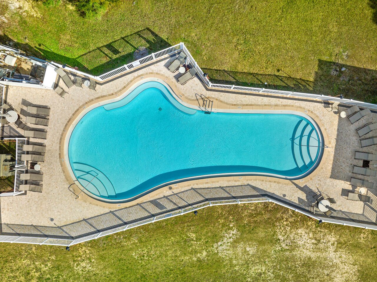 Vista aerea di una piscina dalla forma sinuosa, circondata da una terrazza con lettini e una recinzione.