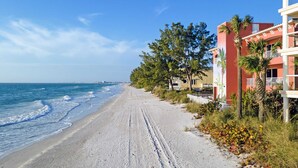 A beach with a clear sky, a sandy path, and a building with balconies and palm trees.