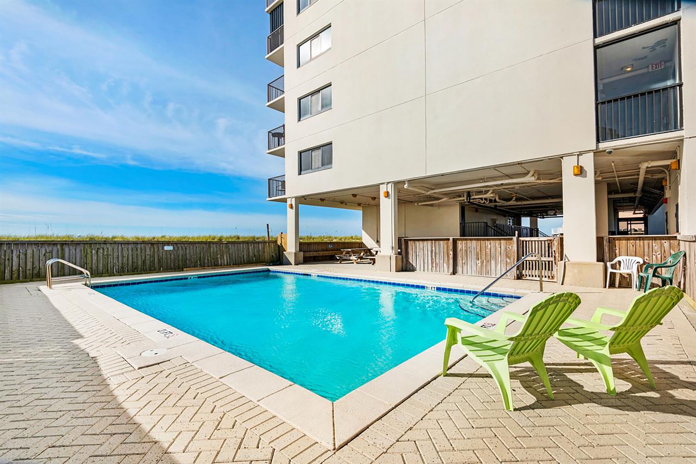 A swimming pool with a clear blue water, surrounded by a brick deck. There are green chairs and a wooden deck area with a view of the ocean.