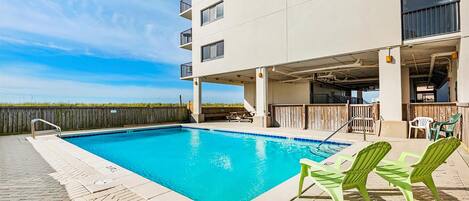 A swimming pool with a clear blue water, surrounded by a brick deck. There are green chairs and a wooden deck area with a view of the ocean.