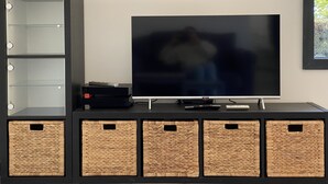 Een flatscreen televisie op een zwarte standaard, met een afstandsbediening, een toetsenbord en een klein kastje. Daaronder staan vijf geweven manden.