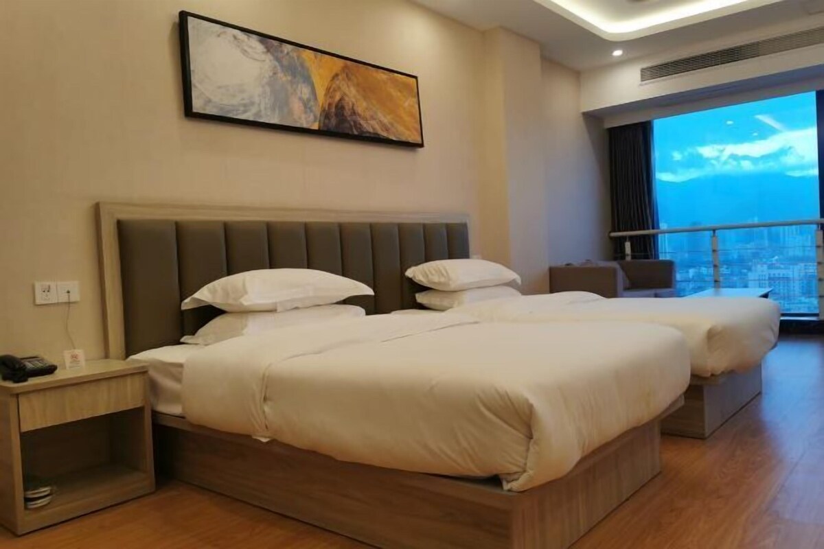 A hotel room with two beds, a nightstand, a painting on the wall, and a large window with a city view.