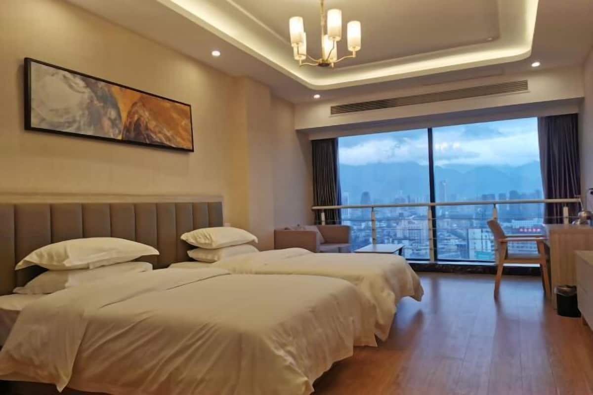 Ein Hotelzimmer mit zwei Betten, einem großen Fenster mit Blick auf die Stadt, einem Gemälde an der Wand und einer Kristallleuchter.