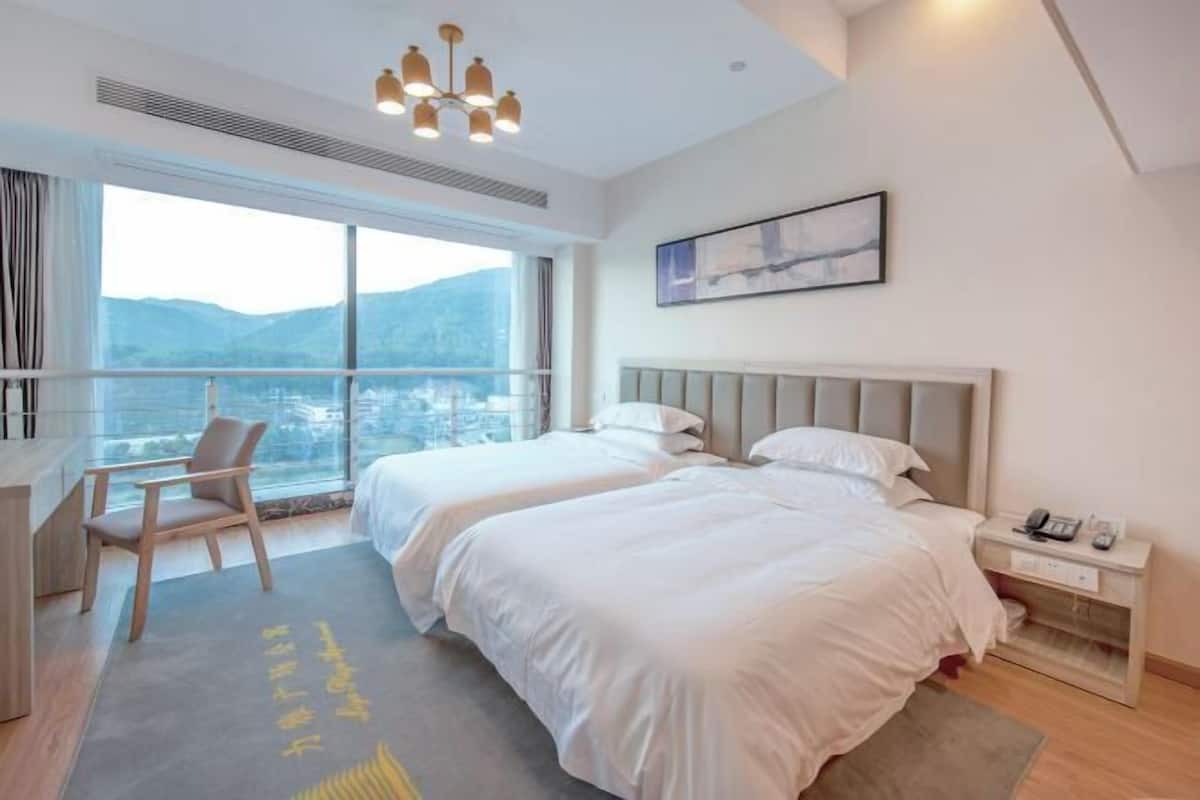 Ein Hotelzimmer mit zwei Betten, einem Schreibtisch, einem Sessel, einem Nachttisch und einem großen Fenster mit Blick auf die Berge.