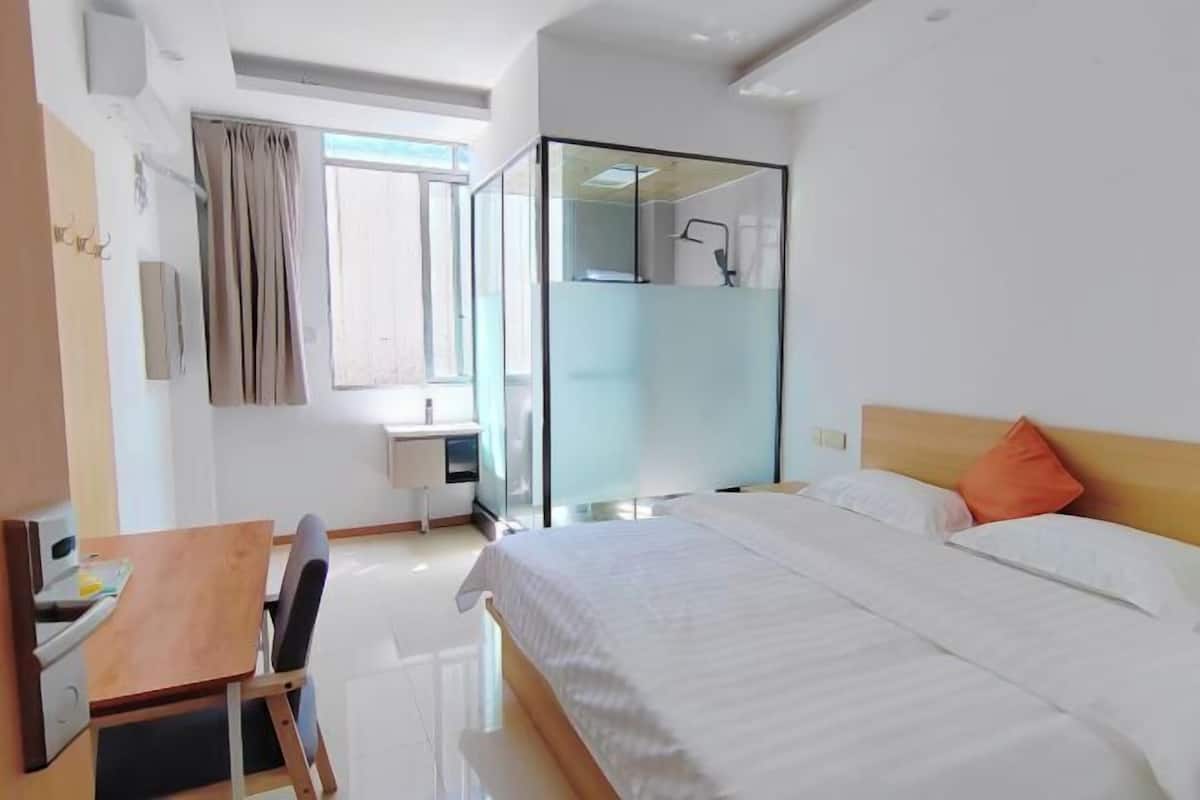 Una habitación de hotel moderna con cama, escritorio, silla y zona de ducha con paredes de vidrio.