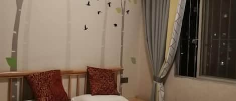 A bedroom with a bed, two pillows, a wall with a tree decal and birds, a window with curtains, and a nightstand.