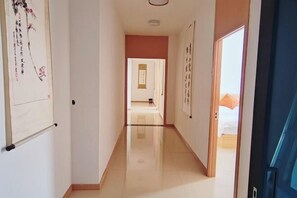 Un corridoio d'hotel con pavimento lucido, una porta a destra e un letto visibile attraverso una porta aperta in fondo.
