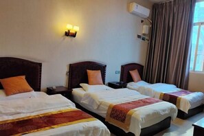 Habitación de hotel con tres camas individuales, cada una con cabecera y mesita de noche. Hay un aire acondicionado de pared y una ventana con cortinas.