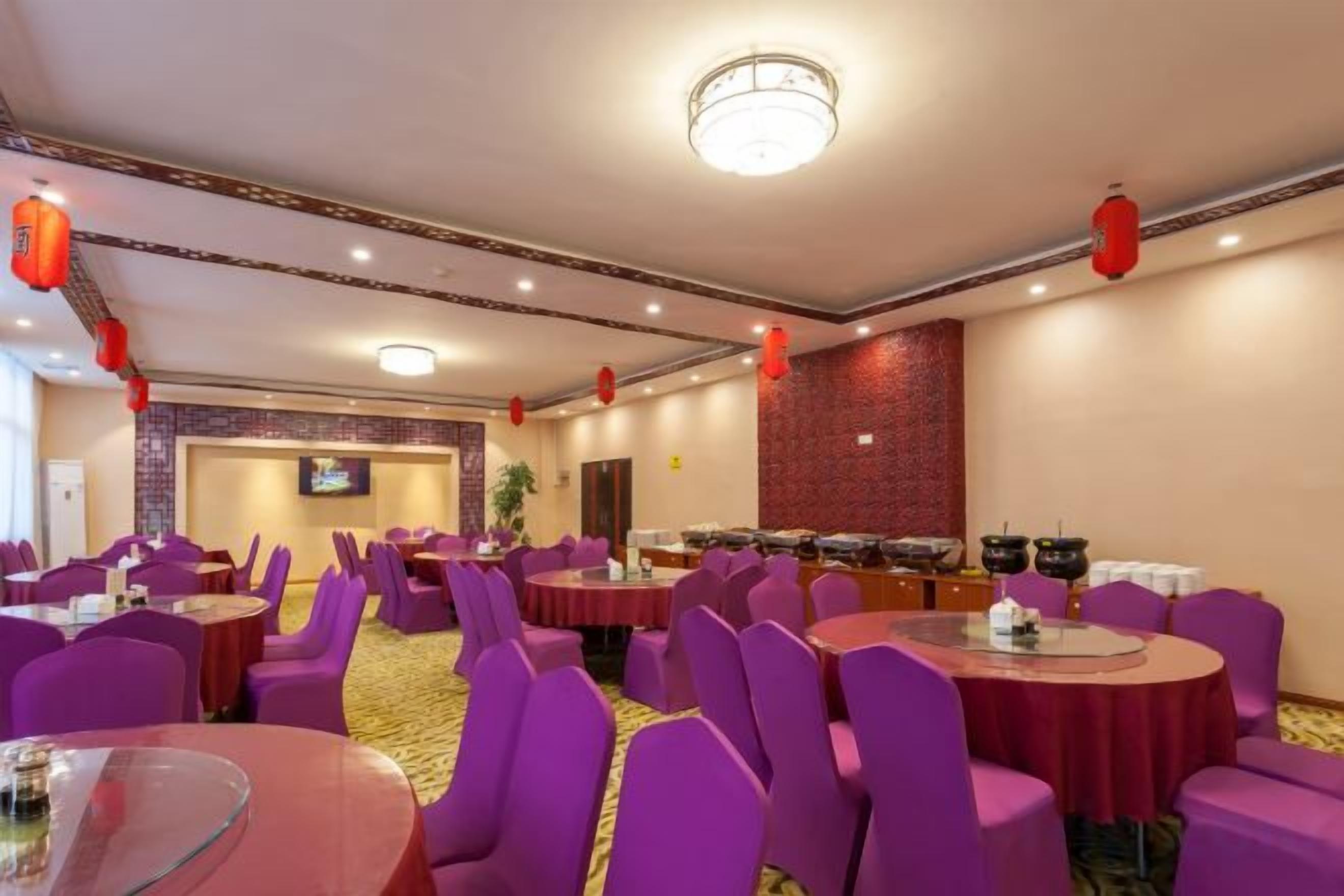 Un salón de banquetes con mesas redondas y sillas moradas, una zona de bufé y faroles rojos en el techo.