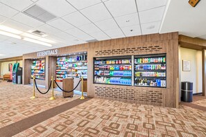 Un vestíbulo de hotel con un quiosco 'ESSENTIALS' repleto de refrigerios y bebidas, un cubo de basura y una alfombra con diseño de madera.