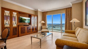 A spacious living room with a large flat-screen TV, a comfortable sofa, a glass coffee table, and a view of a body of water through large windows.