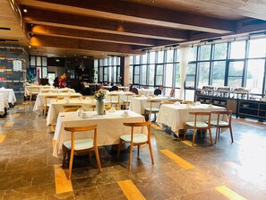 A spacious dining area with multiple tables set for service, large windows providing natural light, and a kitchen area visible in the background. - Blossom Hill Hotel Ningbo Hanling (Ningbo)