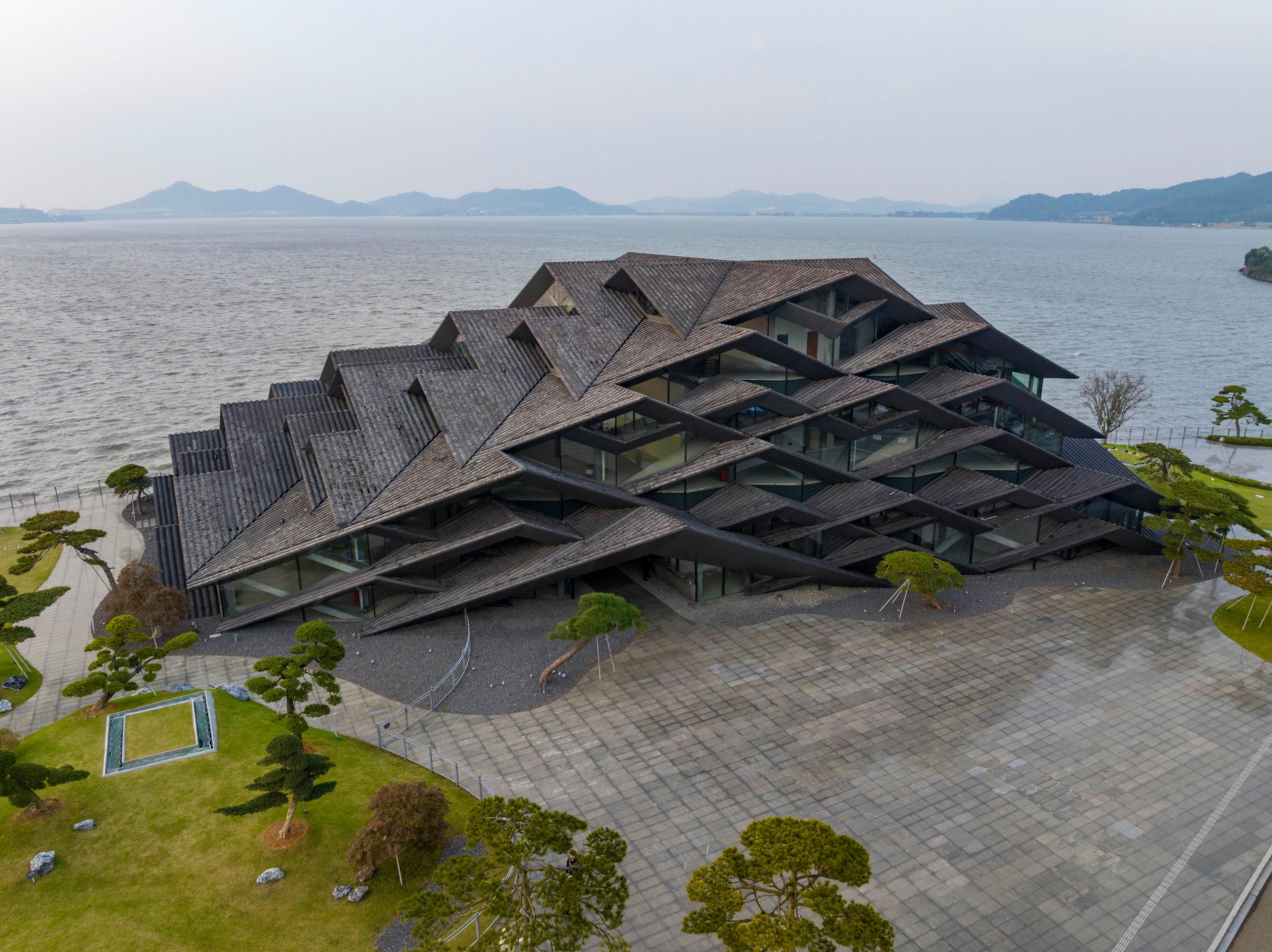A modern building with a unique geometric roof design, situated by a body of water with mountains in the background.