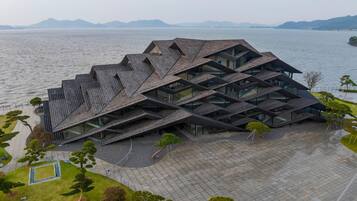 A modern building with a unique geometric roof design, situated by a body of water with mountains in the background.