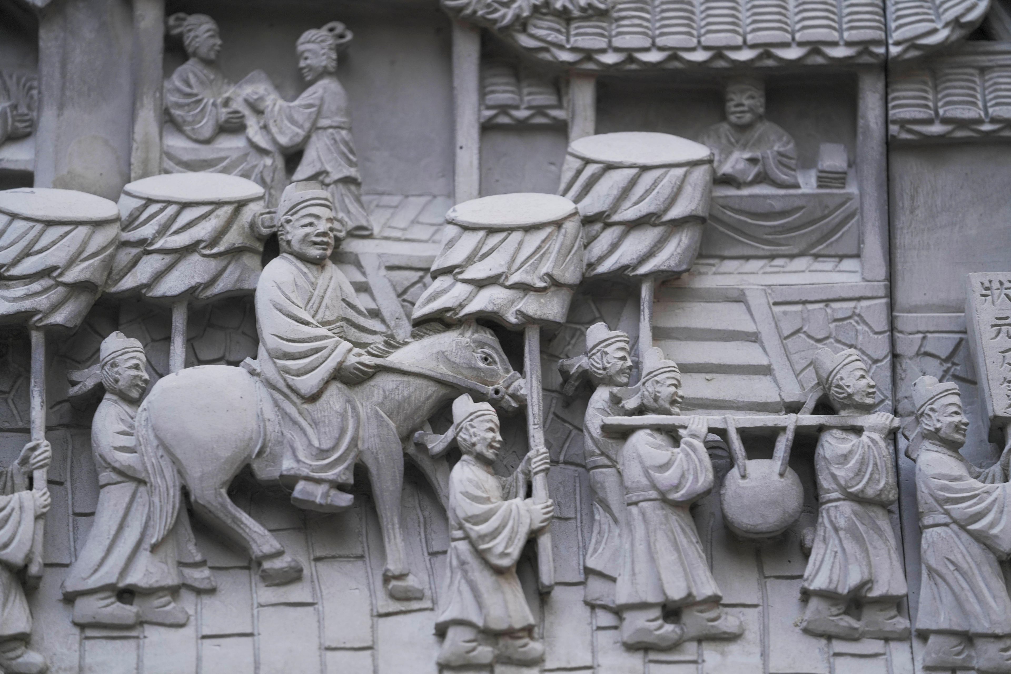 A detailed stone carving of a historical scene with figures, including a seated individual on a horse and others carrying drums and a basket.