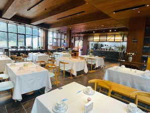 Restaurante com várias mesas e cadeiras, área de cozinha e janelas amplas que permitem entrada de luz natural.