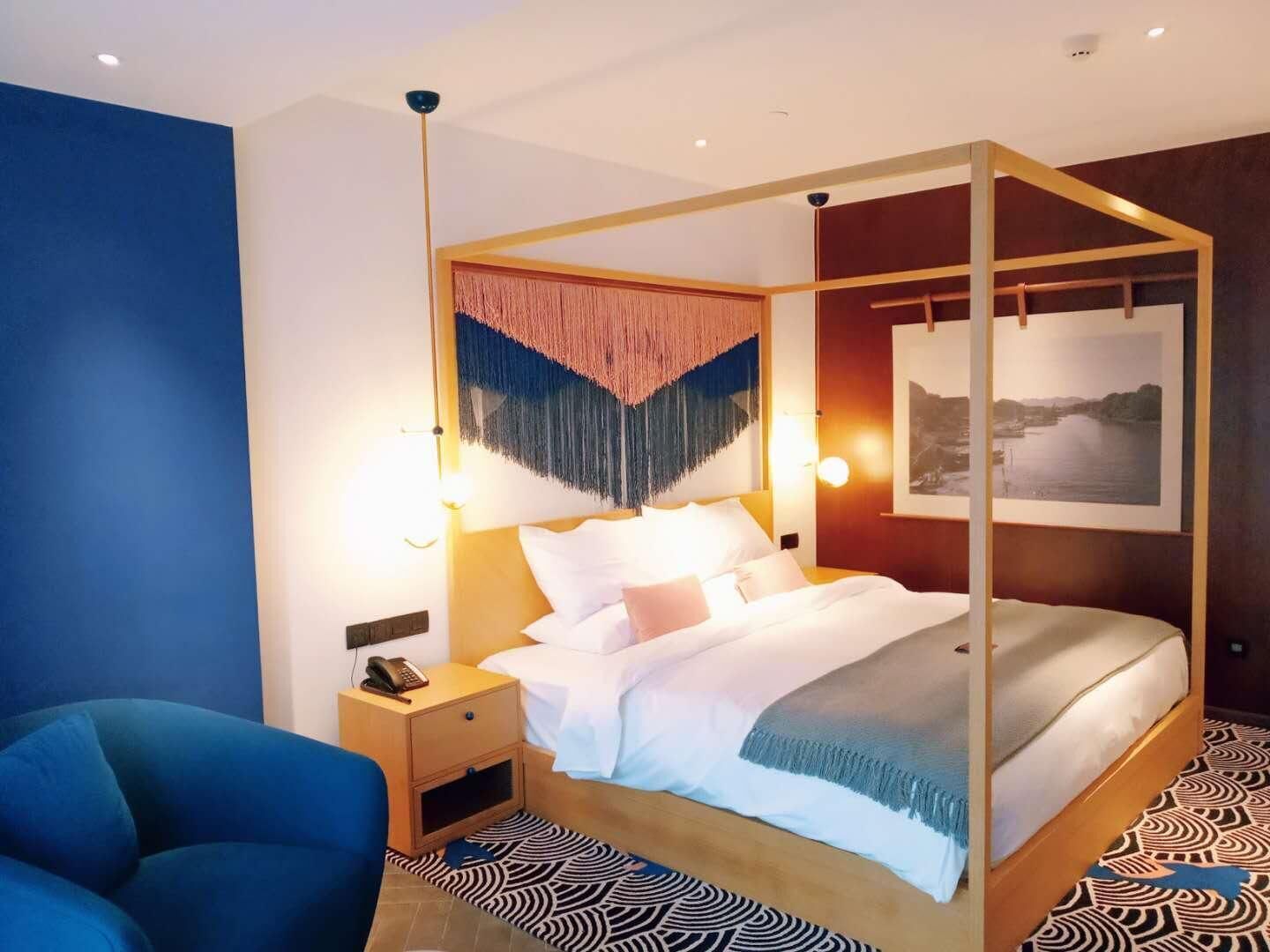 A modern hotel room with a four-poster bed, a nightstand with a phone, a blue armchair, and a patterned rug.