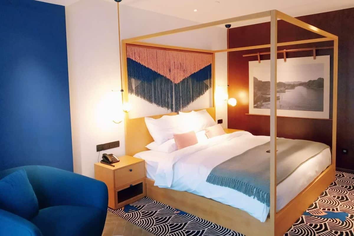 A modern hotel room with a four-poster bed, a nightstand with a phone, a blue armchair, and a patterned rug.