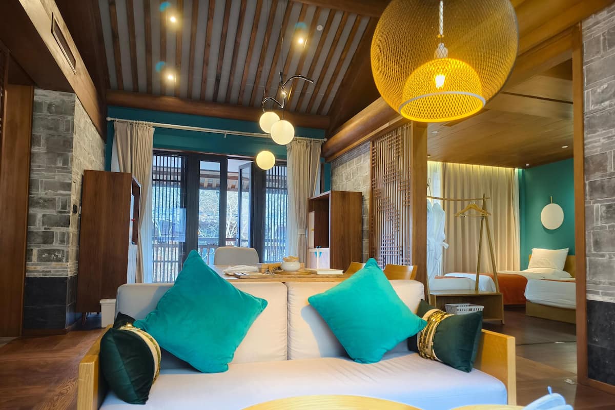 A modern hotel room with a wooden ceiling, a large window, a sofa with teal cushions, and a hanging lamp.