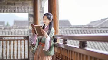 A person in traditional attire reading a book on a balcony with a wooden railing and stone walls in the background.